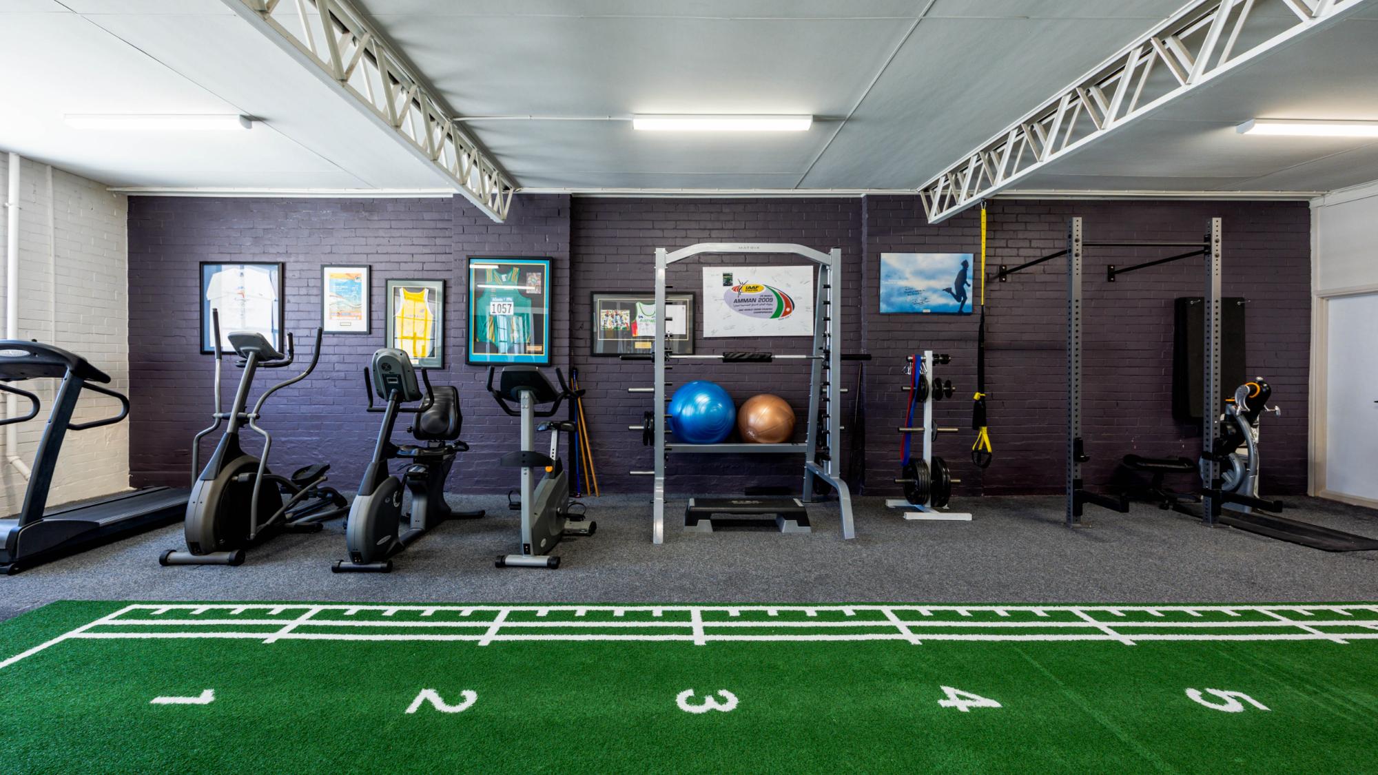 A gym with a sprint or jumping track in the middle, and cardio, strength, and other gym equipment at the back with a dark grey wall and white ceiling.