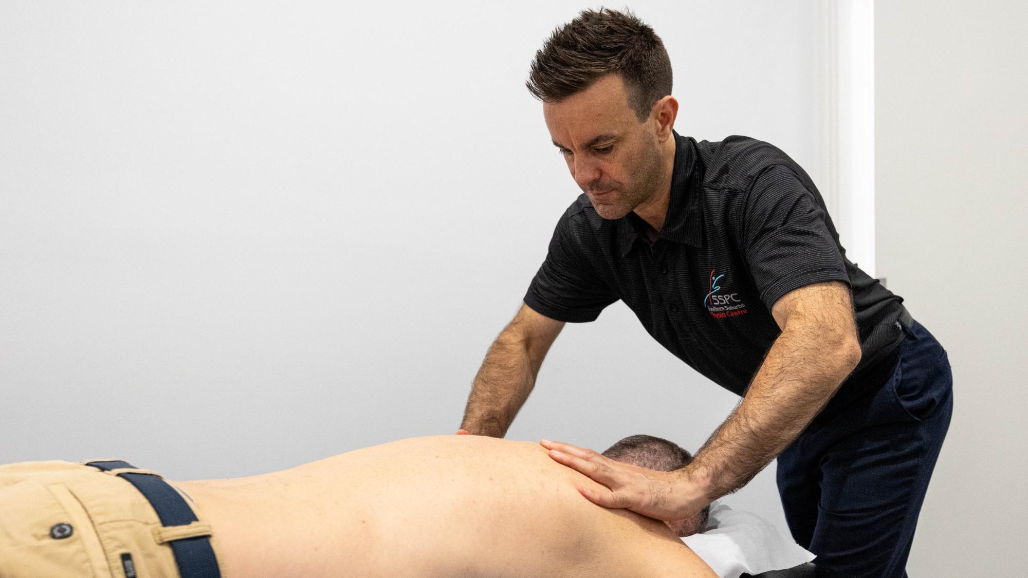 A young man receiving a healing massage from a specialised physiotherapist at a clinic in Bentleigh East, Melbourne. There is a white background and the masseuse is remedying the young man's shoulder pain.